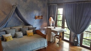 A bedroom with a canopy bed, a bedside table with a lamp, and a view of greenery through the window.