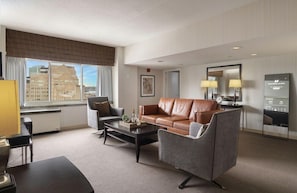 Una habitación de hotel moderna con un sofá de cuero, sillones, una mesita de centro y vistas a la ciudad a través de amplios ventanales.