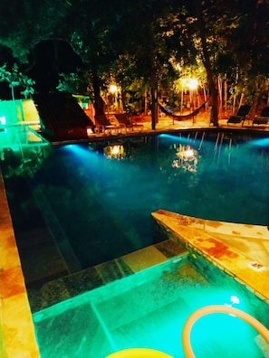 Una piscina nocturna con sillones de descanso y una hamaca, rodeada de árboles e iluminada con luz ambiental.