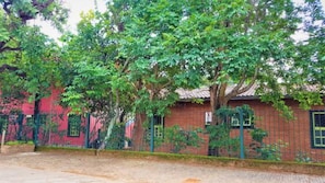 Un edificio de ladrillo rojo con techo de tejas, rodeado de frondosos árboles verdes y una valla de metal.