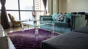 A modern living room with a glass coffee table, a teal sofa with patterned cushions, a purple shag rug, and a city view through large windows. - Pelicanstay Seaview Suites in JBR Walk (Dubai)