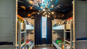 Une chambre avec des lits superposés, une fresque représentant un ciel étoilé au plafond et une fenêtre ornée de rideaux à motifs d’étoiles.