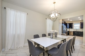 Un coin repas avec une table blanche et des chaises grises, un lustre et une cuisine en arrière-plan.