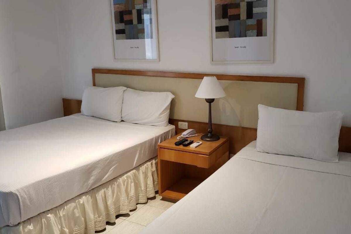 A hotel room with two single beds, a wooden nightstand with a lamp, and two framed pictures on the wall.