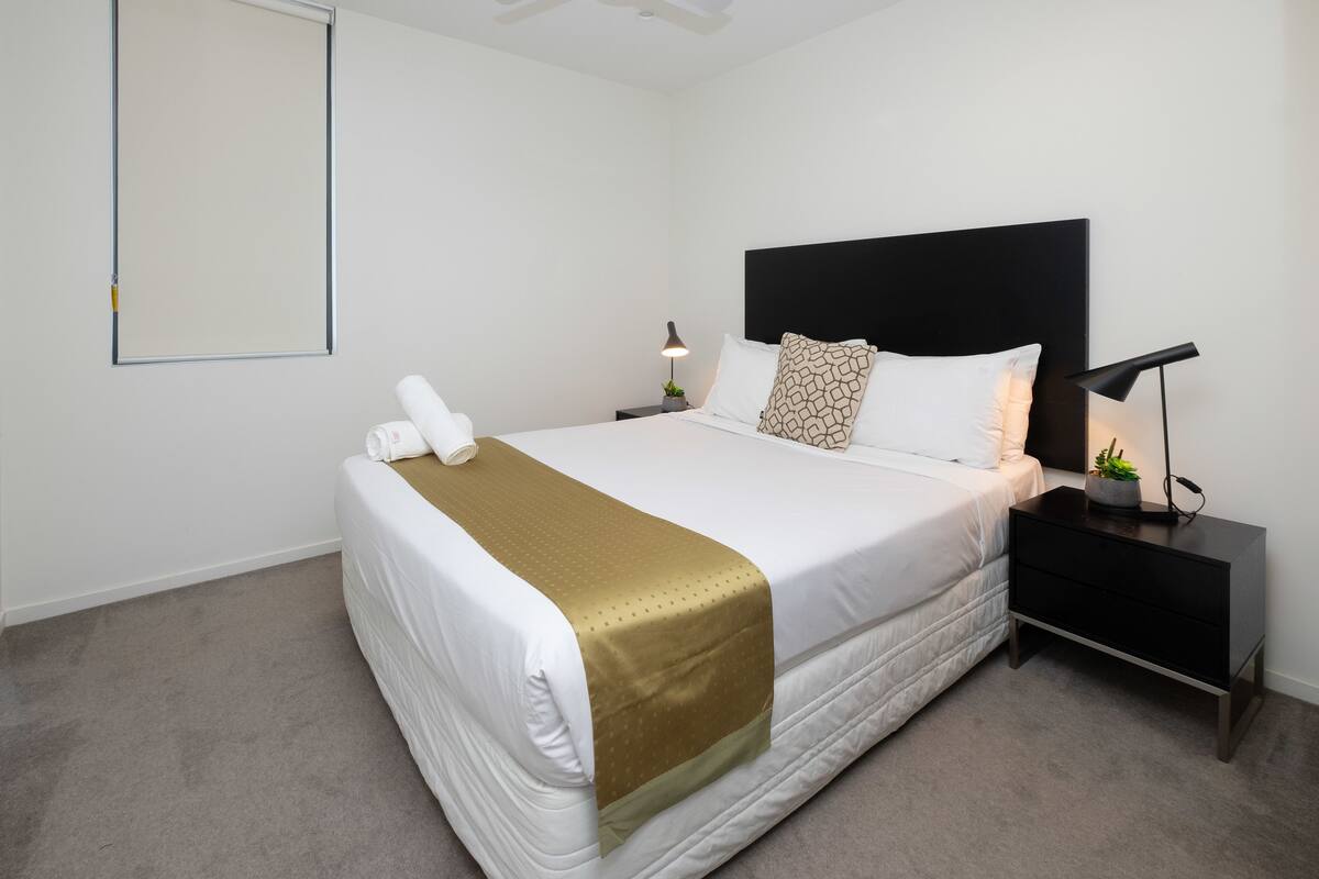 Una cama cuidadosamente tendida con un plaid dorado, almohadas blancas y una mesita de noche negra con una lámpara.