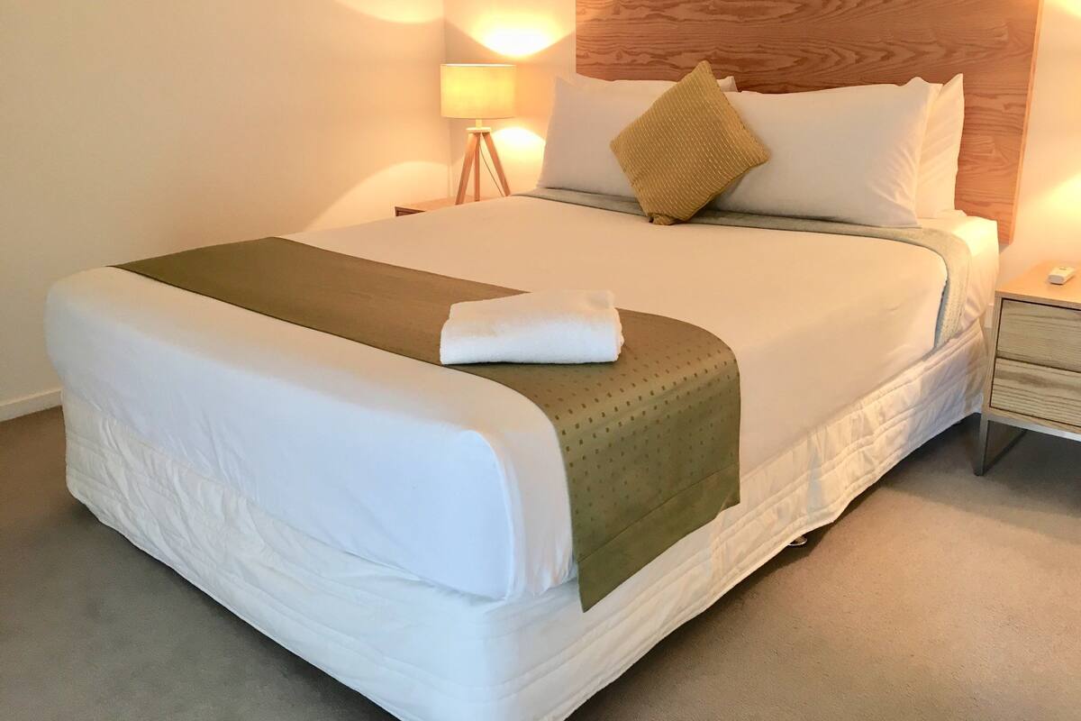 Una cama bien hecha con cabecera de madera, una lámpara de noche y una mesita auxiliar.