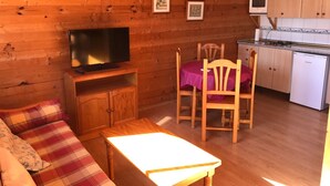 Interior acogedor de una cabaña con techo de madera, un sofá, una mesa de comedor, un televisor y una cocina con armarios y un refrigerador.