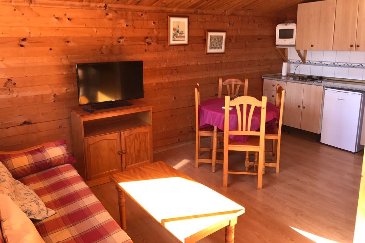 Interior acogedor de una cabaña con techo de madera, un sofá, una mesa de comedor, un televisor y una cocina con armarios y un refrigerador.
