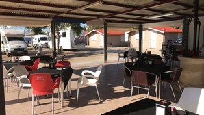 Un café al aire libre con mesas y sillas, una zona cubierta y vistas a la calle con vehículos estacionados.