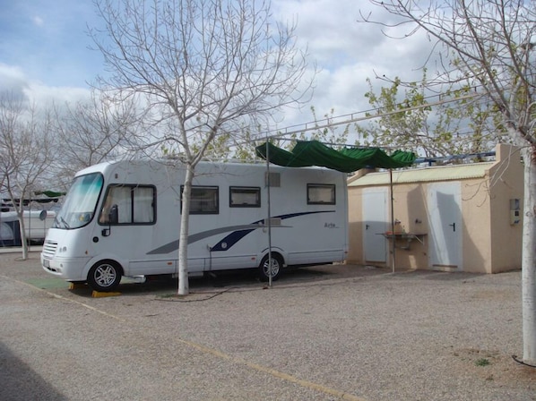 Una caravana blanca estacionada en un estacionamiento, con un toldo verde, árboles y un edificio al fondo.