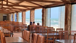 Un restaurante frente al mar con mesas y sillas de madera, amplios ventanales con vistas al océano y un techo de lámina corrugada.