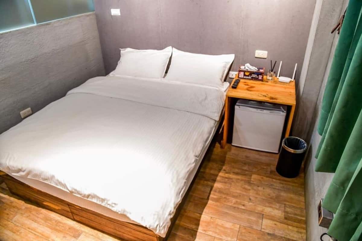 A small, neatly made bed with white linens in a compact room with a wooden floor and a small desk with a lamp and a box of tissues.