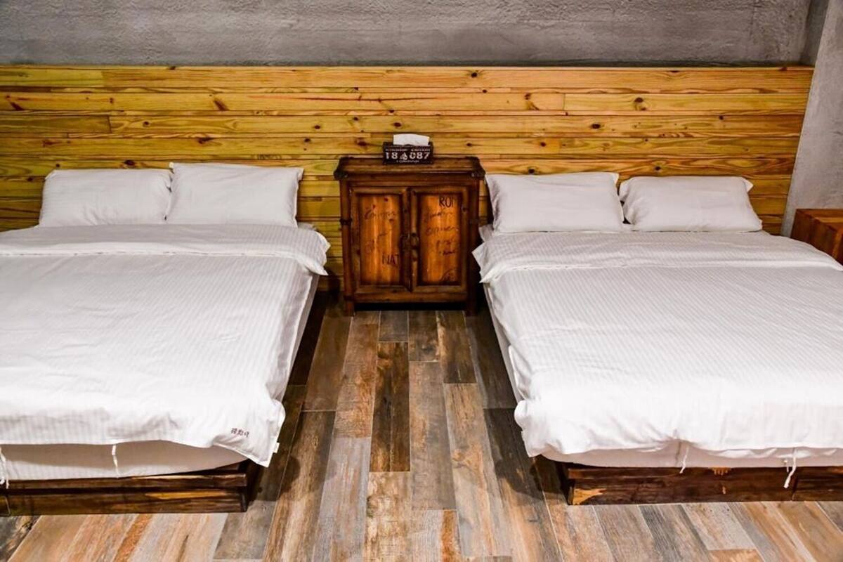 A room with two beds, a wooden headboard, a small wooden cabinet, and a tiled floor.