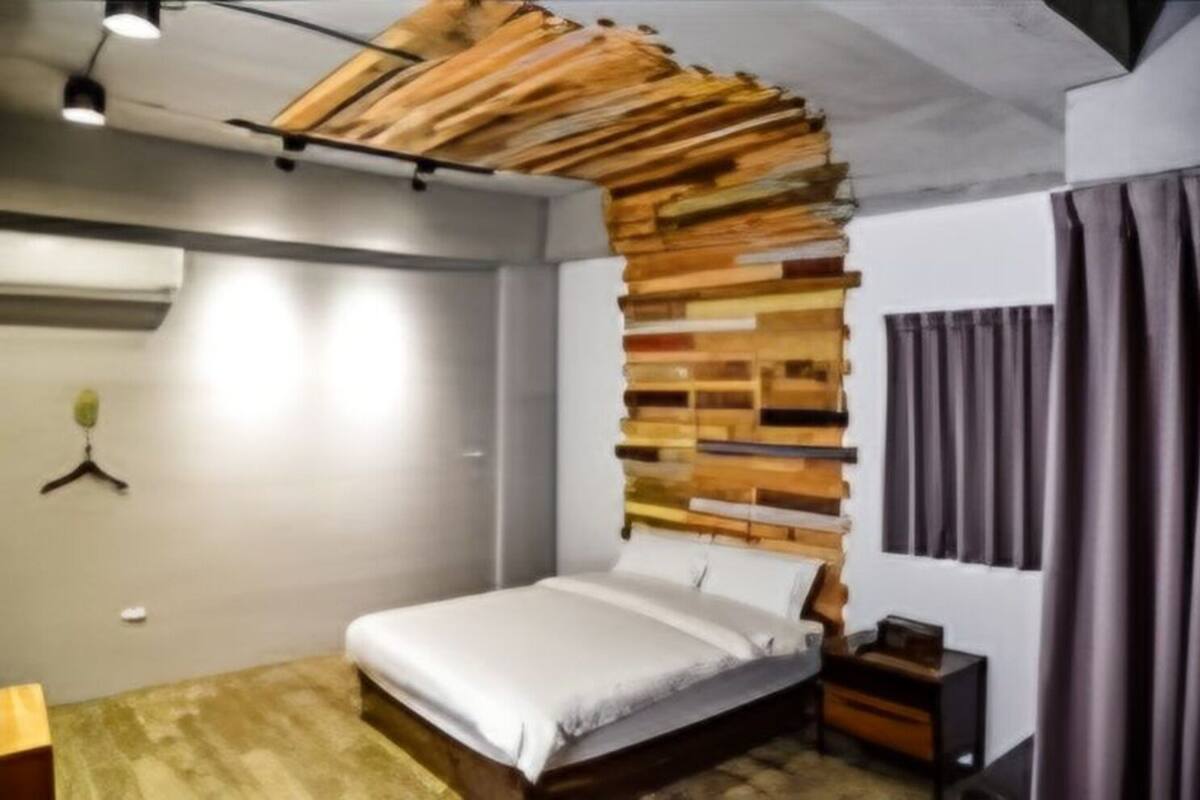 A bedroom with a bed, a wooden headboard, a nightstand, a hanging plant, and a window with curtains.