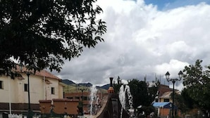 A public square with a central fountain, paved with stone tiles, surrounded by benches and trees.