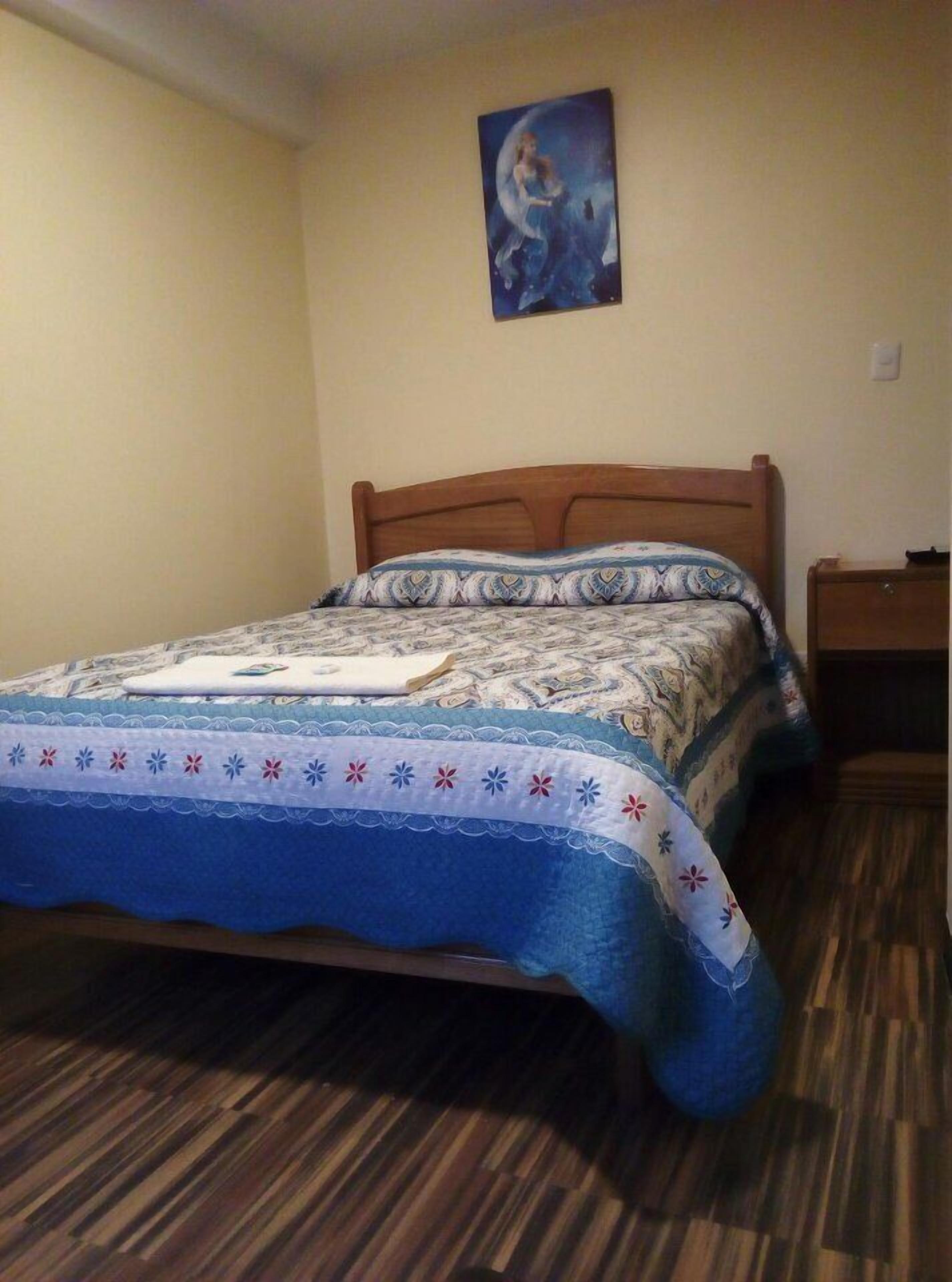 Un dormitorio con una cama de madera, ropa de cama estampada, un cuadro en la pared y una mesita de noche.