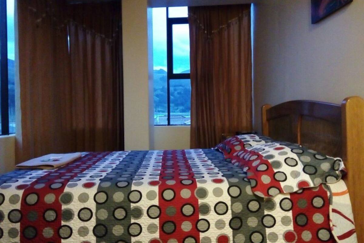 Une chambre à coucher avec un lit recouvert d’une couette à motifs pois, une fenêtre avec des rideaux et une vue sur un paysage montagneux.