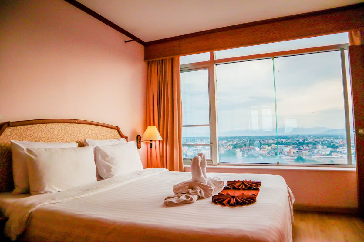 Quarto de hotel com uma cama grande, vista para a cidade pela janela e um abajur na parede.