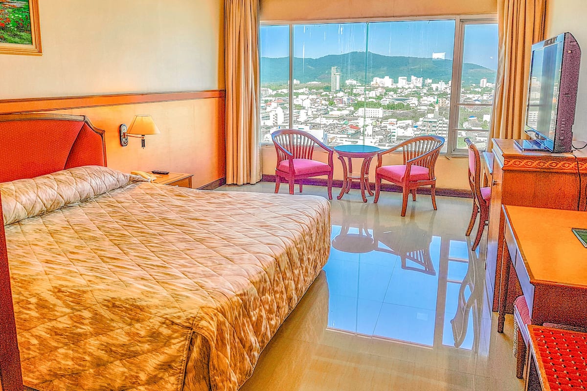 Quarto de hotel com uma cama grande, uma mesa com cadeira, televisão e vista para o horizonte urbano através de uma janela ampla.