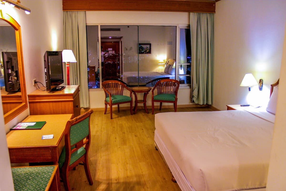Quarto de hotel com cama, escrivaninha com computador, duas cadeiras, janela com cortinas e mesa de cabeceira com abajur.