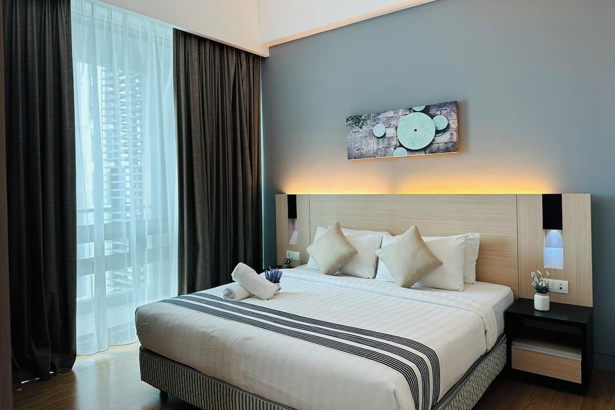Quarto de hotel moderno com uma cama grande, cabeceira com iluminação integrada, mesa de cabeceira com abajur e vista para edifícios através de cortinas finas.