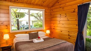Habitación de cabaña de madera con una cama, dos lámparas de noche, una ventana que ofrece vistas a un coche y follaje, y una cortina a la derecha.