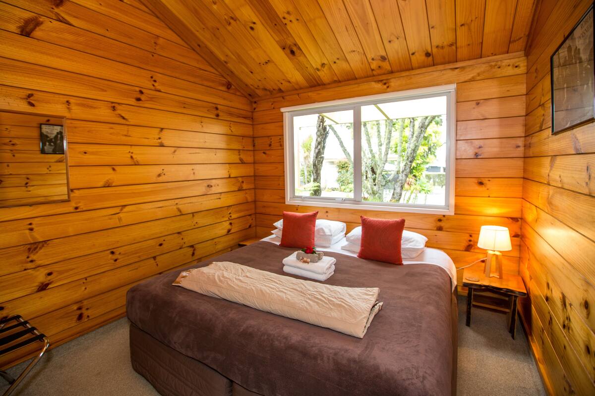 Una habitación de cabaña de madera con una cama, almohadas, una mesita de noche y una ventana.