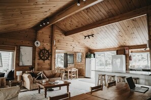 A cabin interior with a wooden ceiling, a dining area, a kitchen with a refrigerator, and a large window. - Tatahi Cove - Hostel (Hahei)