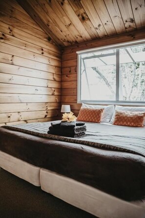 A wooden cabin bedroom with a bed, pillows, and a window. - Tatahi Cove - Hostel (Hahei)