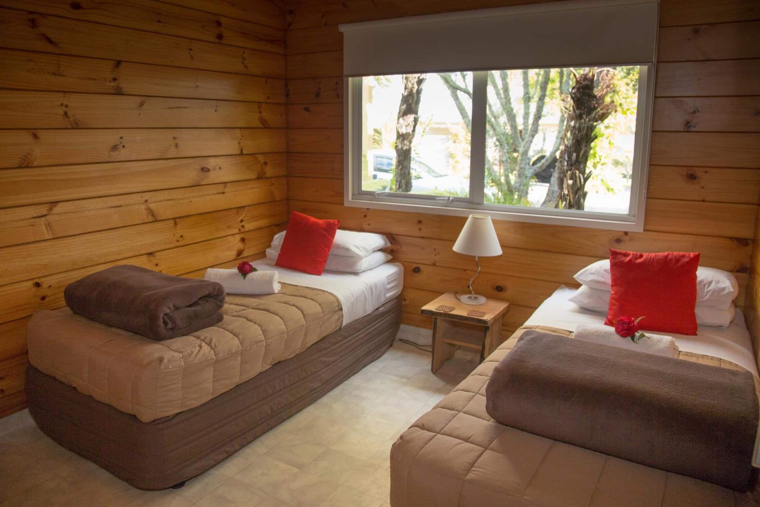 A cozy cabin-style room with two beds, a window with a view of trees, and a small table with a lamp.