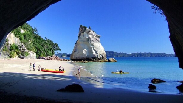 Una playa con una gran formación rocosa, kayaks y gente en la arena.