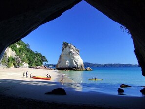 A beach with a large rock formation, kayaks, and people on the sand. - Tatahi Cove - Hostel (Hahei)