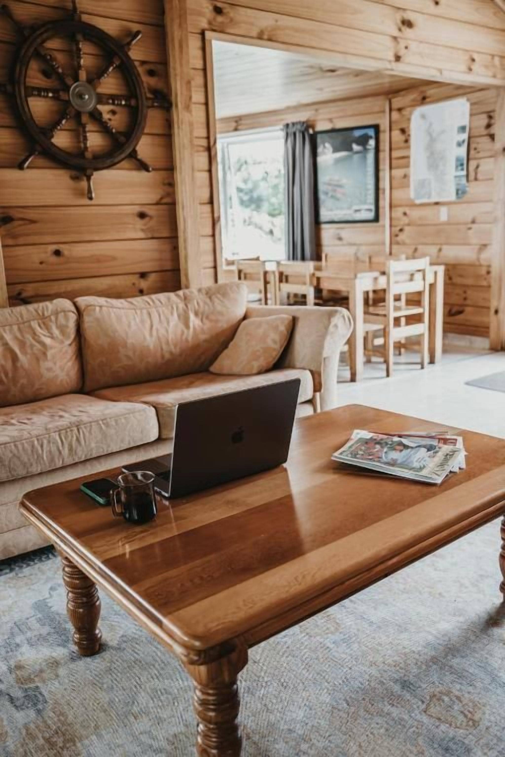 Un intérieur en bois avec un canapé, une table basse en bois sur laquelle se trouvent un ordinateur portable et un magazine, et une fenêtre avec des rideaux.