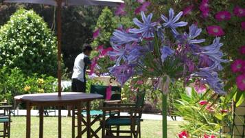 An outdoor seating area with a table and chairs under a white umbrella, surrounded by lush greenery and flowering plants.