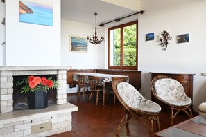 A dining area with a fireplace, wicker chairs, a table with a floral tablecloth, and a window with a view of greenery.