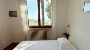 Un dormitorio con cama, ventana con vistas, radiador y una mesita de noche.