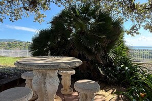A stone table with four stools, surrounded by lush greenery and a view of a distant landscape.