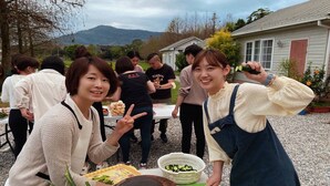 Un grupo de personas reunidas alrededor de una mesa con comida y verduras, al aire libre, con una casa y montañas al fondo.