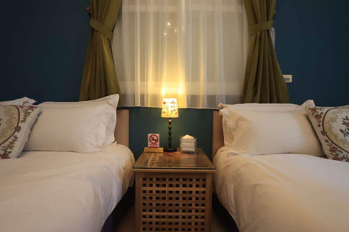 Quarto de hotel com duas camas, uma mesa de cabeceira com abajur e uma janela com cortinas finas.