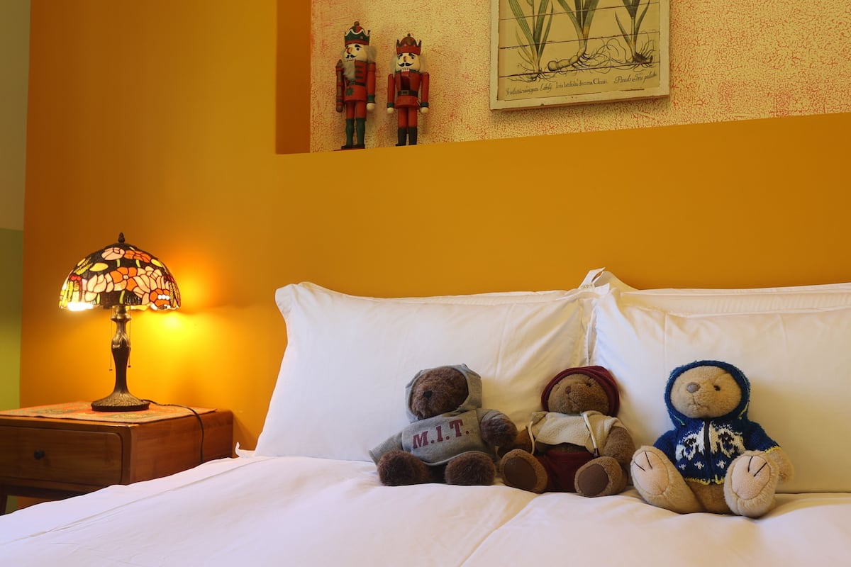 Cama com lençóis brancos, luminária de cabeceira e três ursinhos de pelúcia.