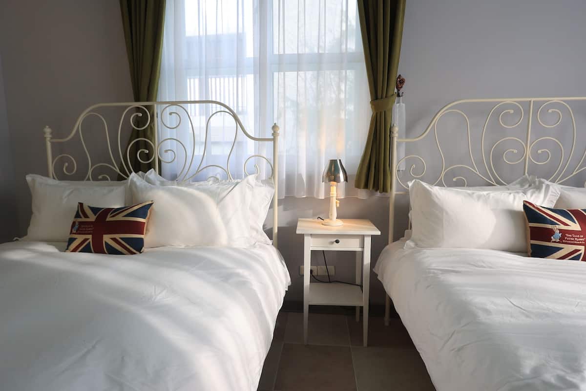 Quarto de hotel com duas camas, cada uma com colcha e almofadas brancas, incluindo uma com padrão da Union Jack. Há uma mesa de cabeceira com abajur e uma gaveta pequena.