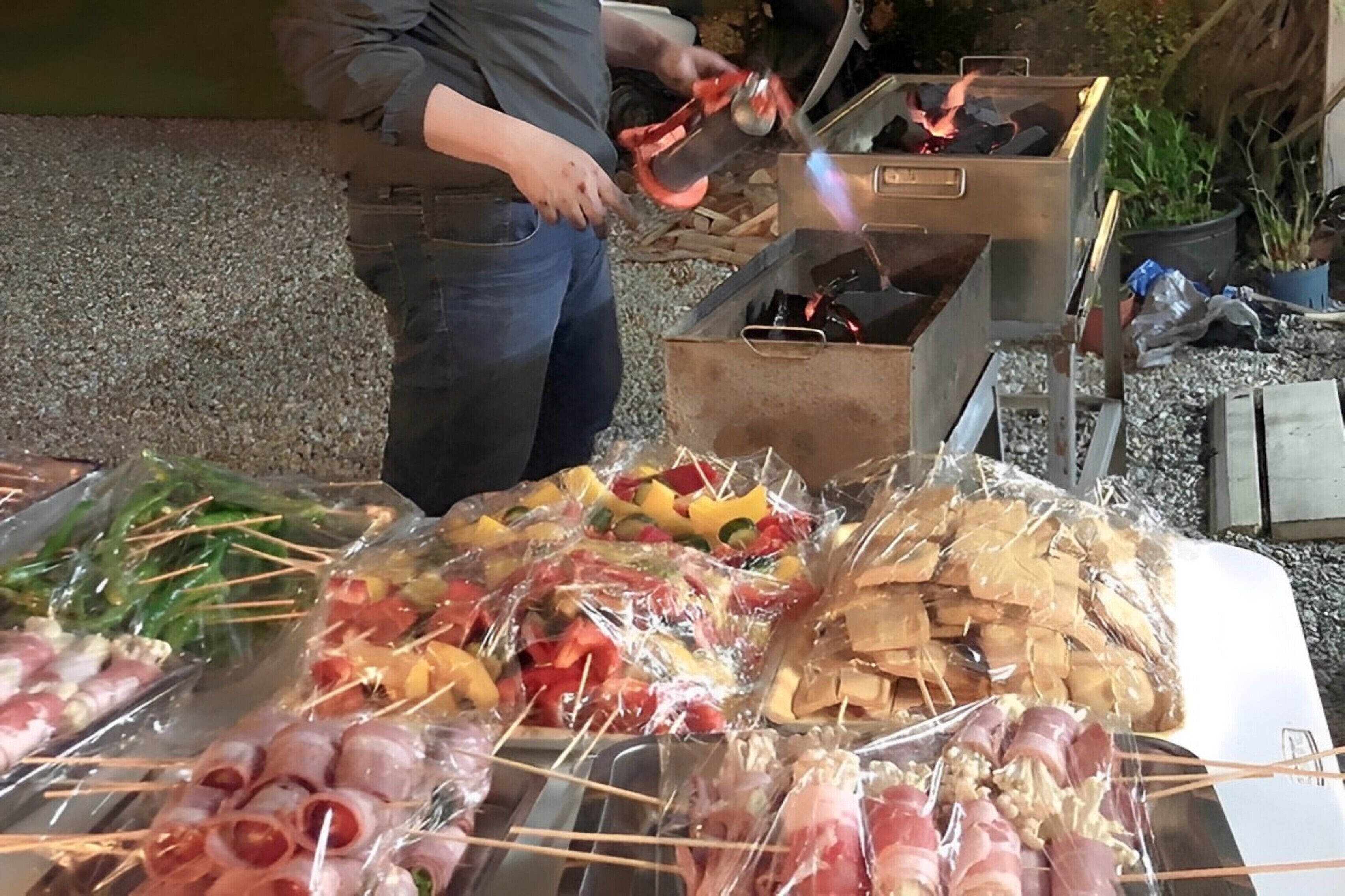 Iemand bereidt eten buiten, met een barbecue en diverse spiezen met vlees en groenten.