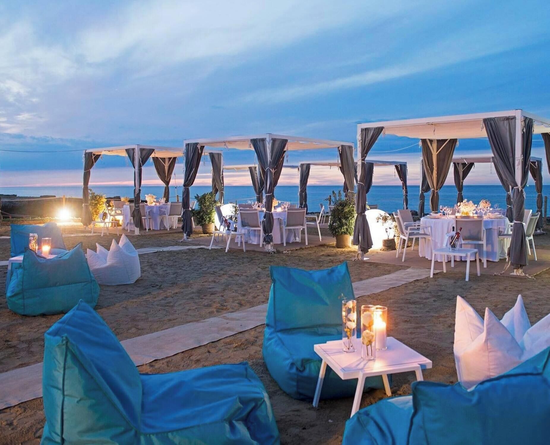 An outdoor dining area with tables and chairs, set against a beach backdrop.