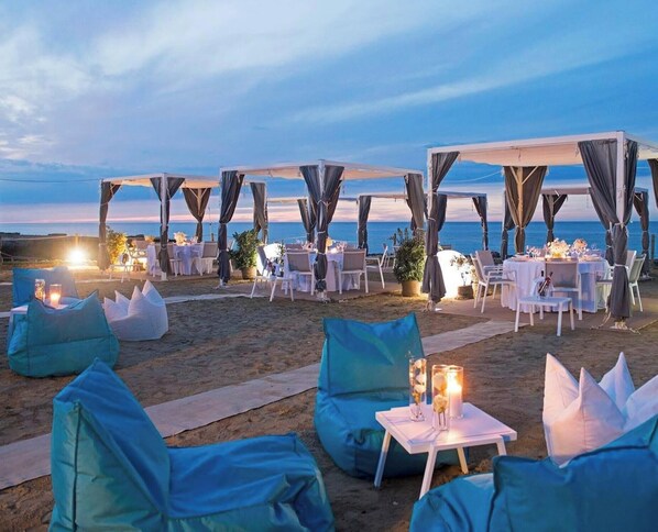 An outdoor dining area with tables and chairs, set against a beach backdrop.