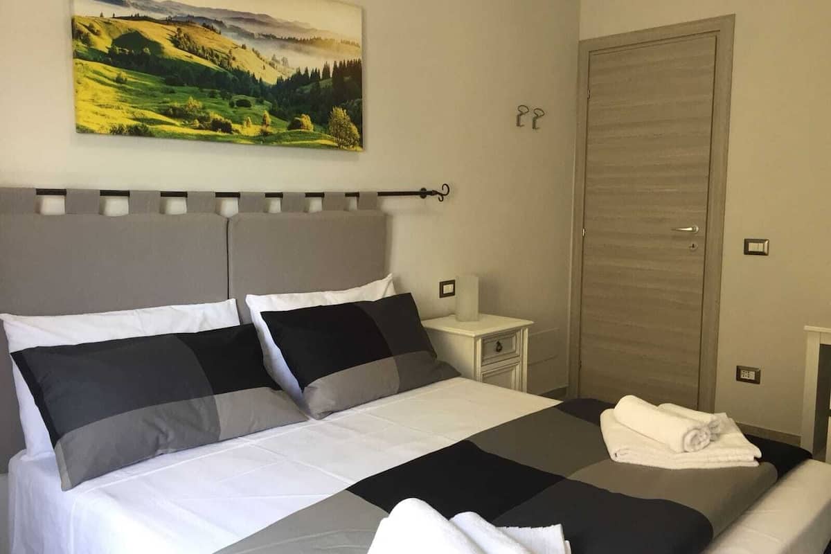 A neatly made bed with white and grey bedding, a nightstand with a lamp, and a painting of a landscape on the wall.