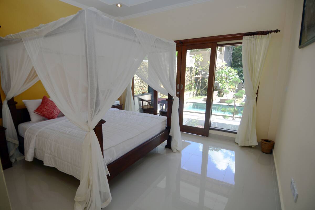 A bedroom with a four-poster bed, a red pillow, and a view of a pool through a glass door.