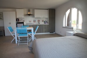 A compact room with a bed, a dining table with chairs, a kitchen area, and a large arched window.