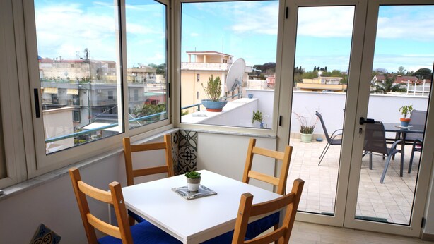 A dining area with a white table and blue cushioned chairs, a potted plant on the table, and a view of the city through large windows. - Nolana 86 (Pompei)