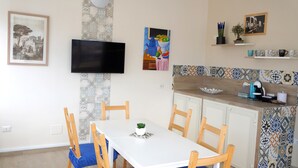 A dining area with a white table and chairs, a TV mounted on the wall, and a kitchen area with a tiled backsplash. - Nolana 86 (Pompei)