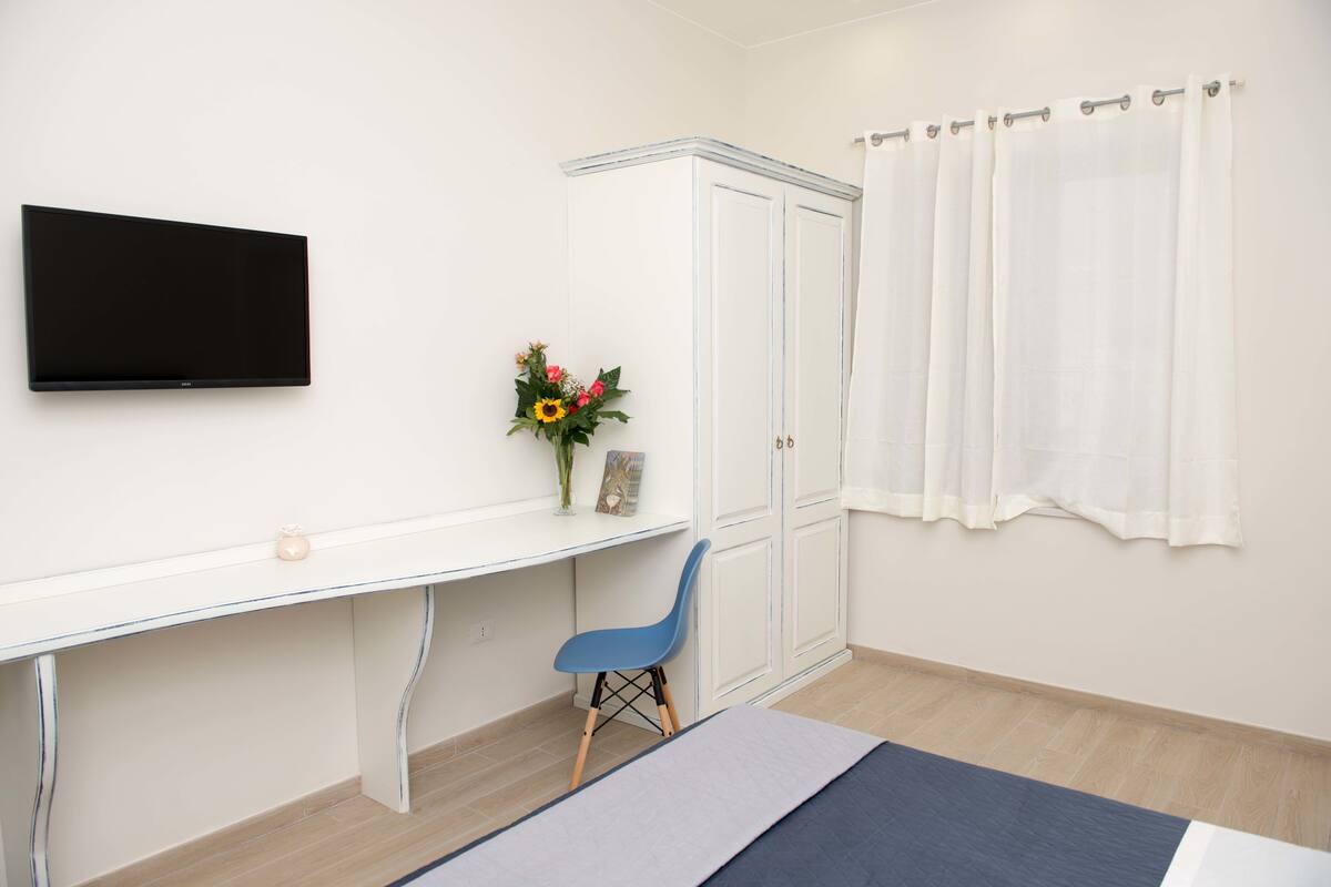 Een kamer met een wandgemonteerde flatscreen-tv, een wit bureau met een bloevas, een blauwe stoel en een bed met een blauw-witte dekbedovertrek.